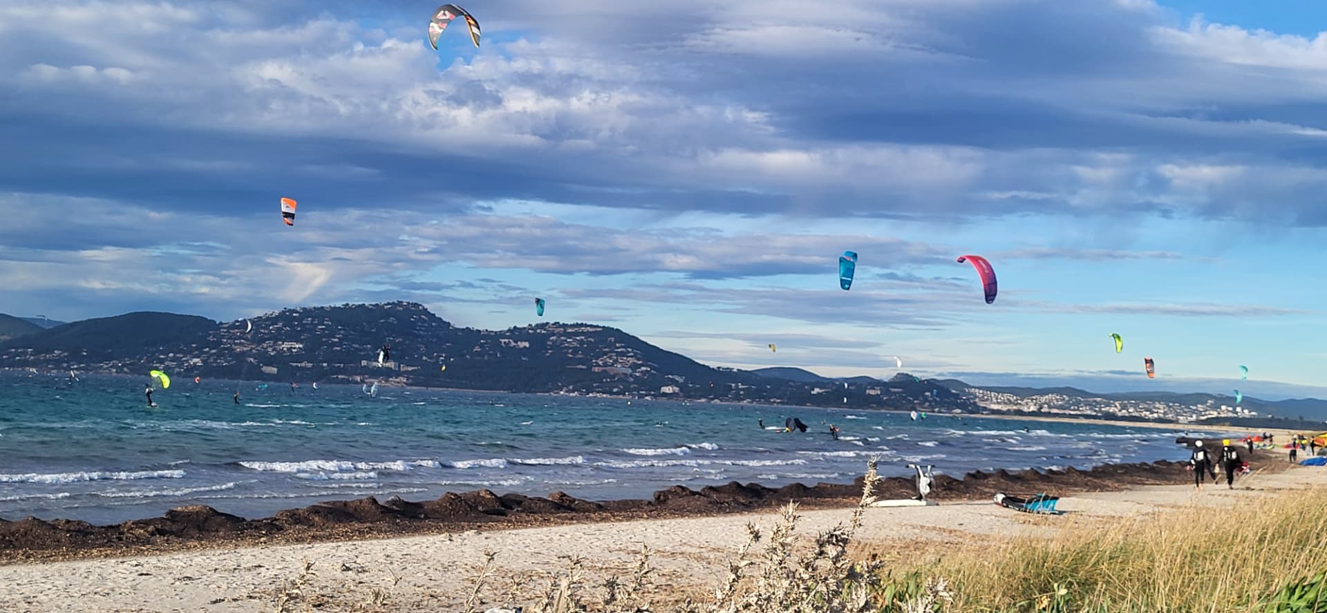 Kitesurfers with colorful sails over the turquoise water at Almanarre, Giens peninsula, with hills along the Var coast