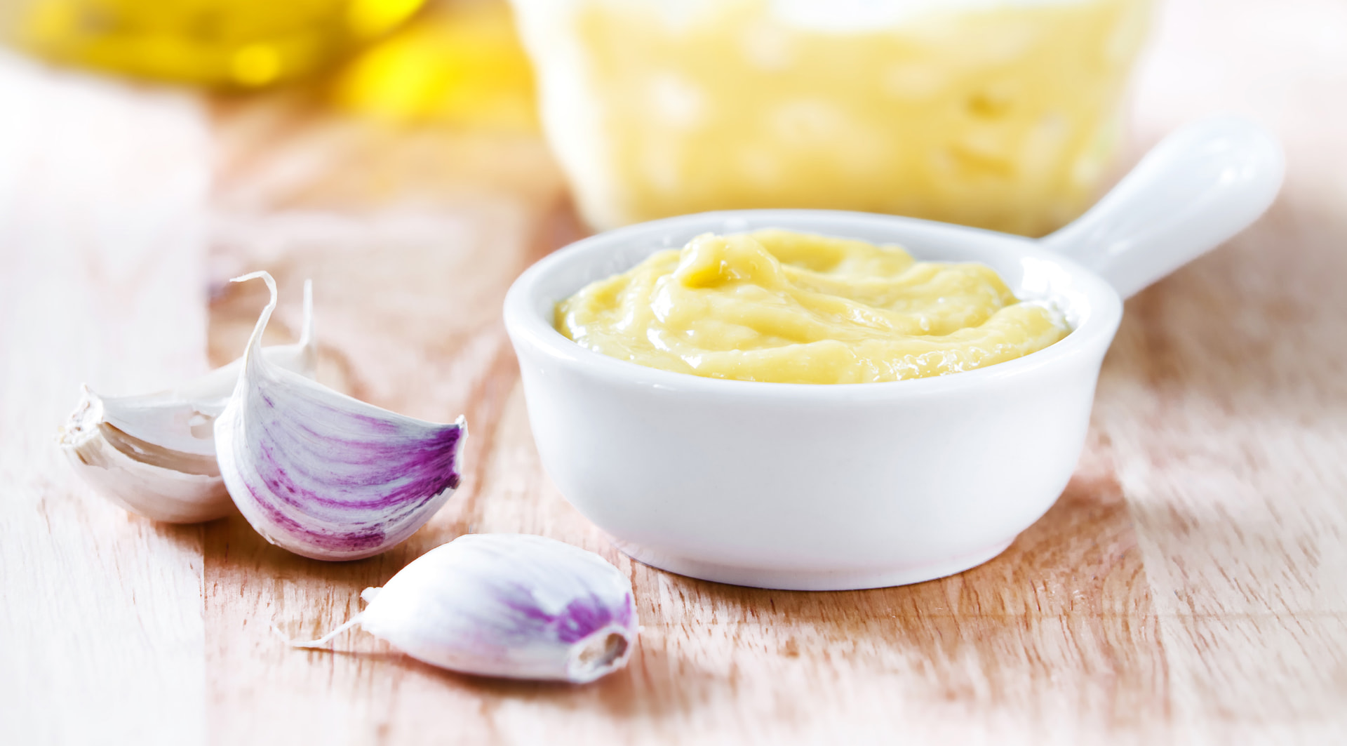 Bowl of golden aïoli with garlic cloves and olive oil alongside