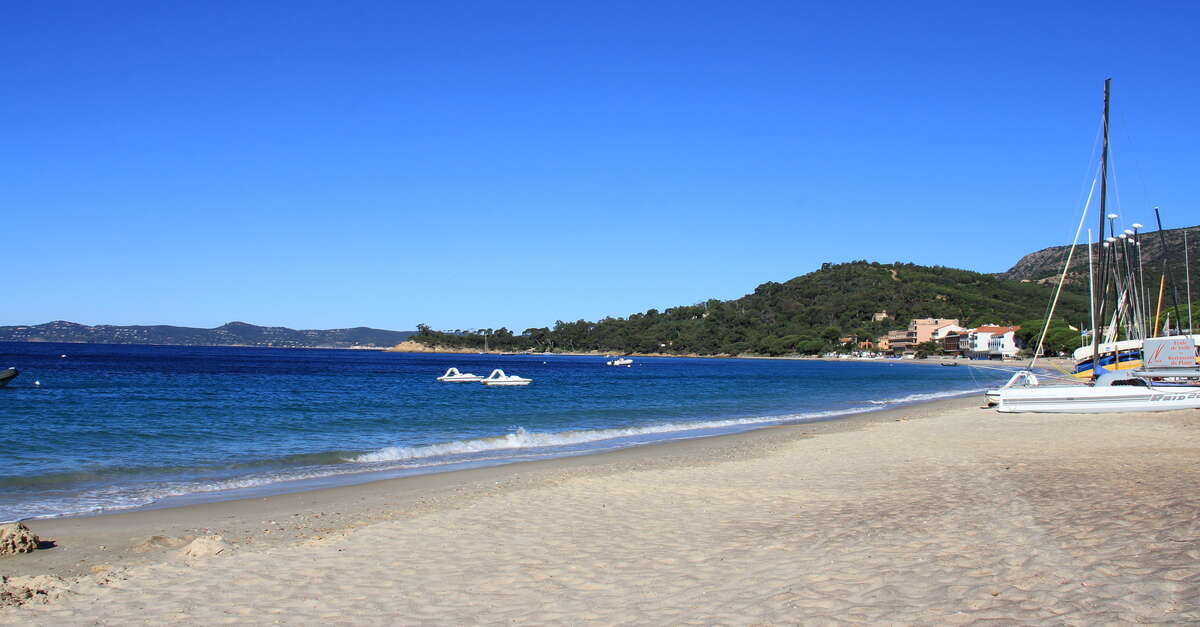 Wide sandy beach at Cavaliere, Le Lavandou, with calm blue water, moored catamarans and the wooded Massif des Maures headland beyond