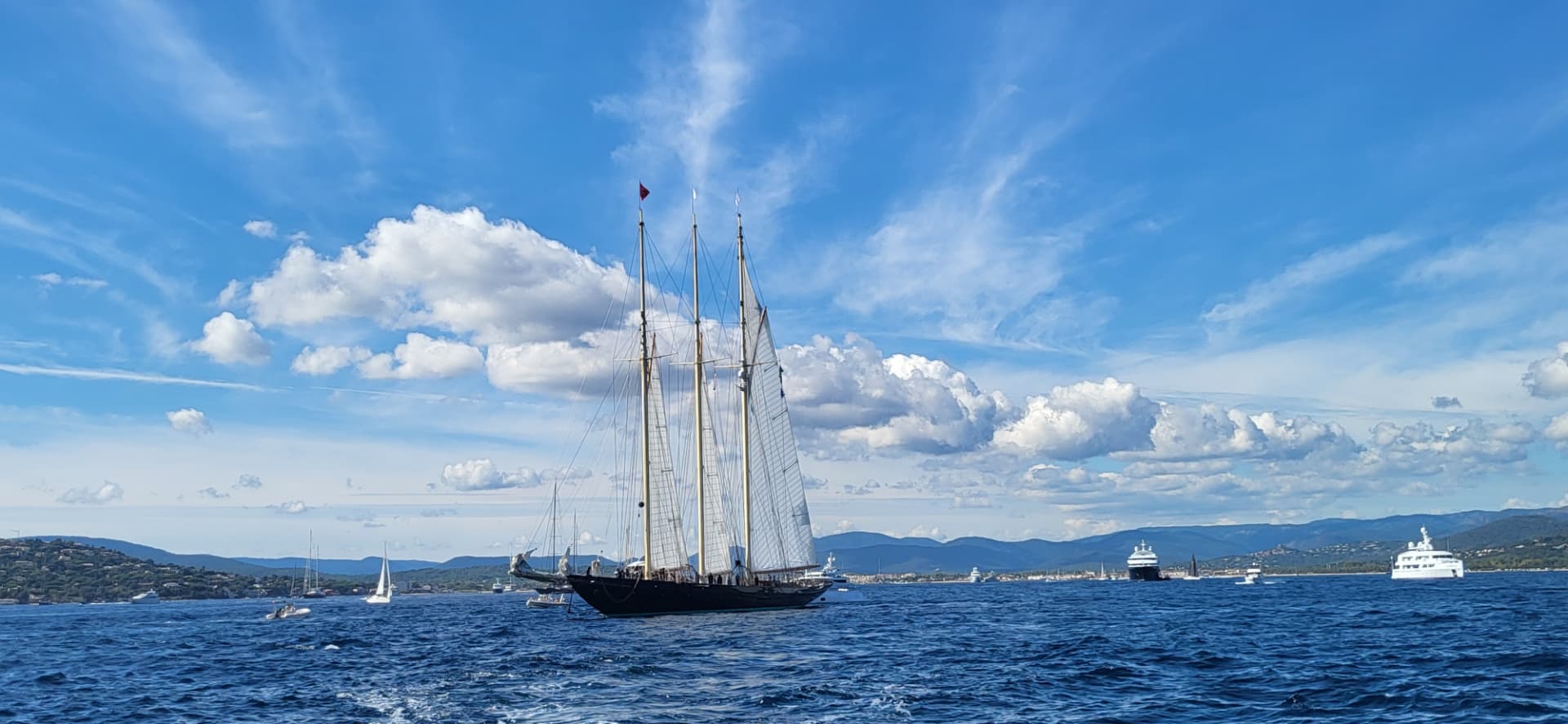 Classic three-masted schooner and yachts sailing in the Gulf of Saint-Tropez under a bright blue sky with coastal hills behind